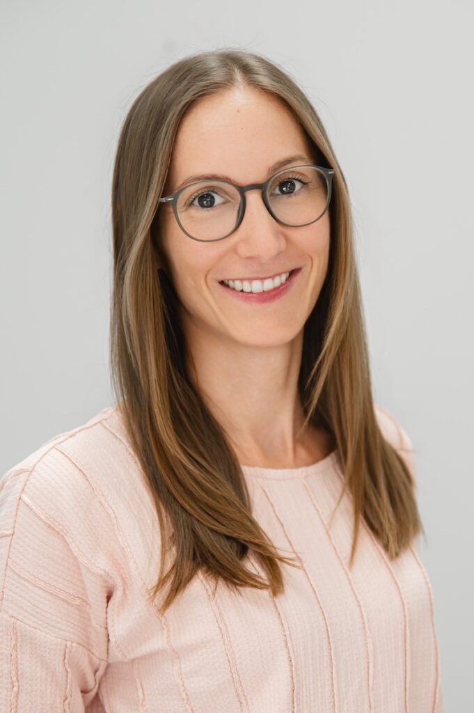 Romana Riener, Therapeutin im Vitanium Therapiezentrum Pro, mit langen braunen Haaren und Brille in einem hellrosa Pullover vor hellem Hintergrund