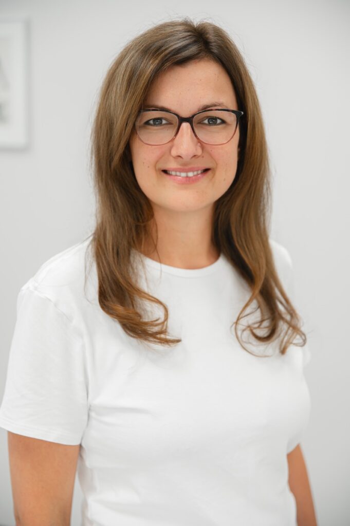 Katharina Stellnberger, Ergotherapeutin im Therapiezentrum Pro, mit Brille und braunen Haaren in weißem T-Shirt, lächelst freundlich in die Kamera.