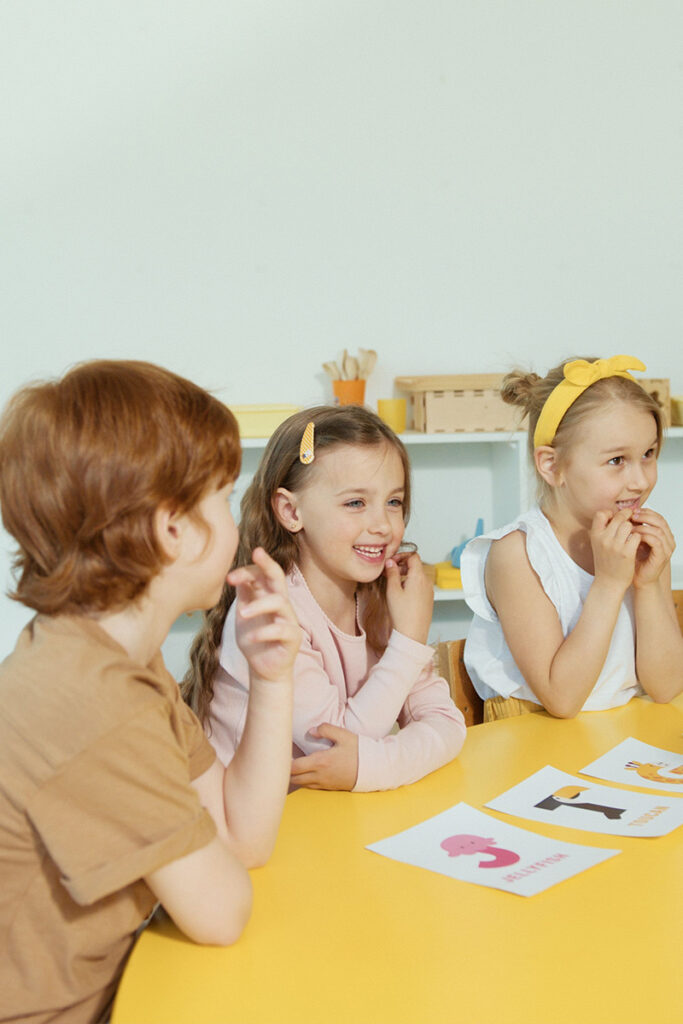 Drei Kinder sitzen in einer hellen Praxis- oder Lernumgebung an einem Tisch. Sie betrachten Bildkarten mit Tieren und wirken aufmerksam und interessiert während einer logopädischen Übung.