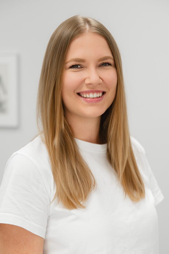 Portraitfoto: Therapeutin Jaqualine Pfarrhofer mit langen blonden Haaren in weißem T-Shirt, lächelt freundlich in die Kamera.