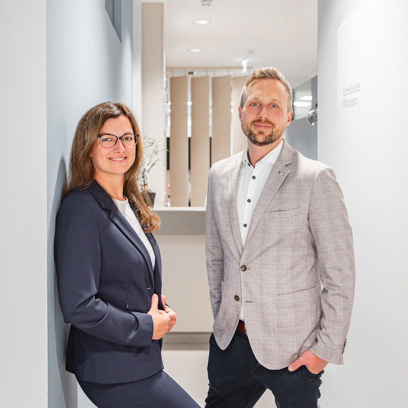 Florian und Katharina stehen in einem hellen Gang der Praxis. Beide tragen Business-Kleidung und blicken freundlich in die Kamera.